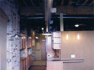 In this old building with timber framed ceilings the galley kitchen in this 600sf loft apartment has modern cabinets tucked into old doorways in the brick wall. Note the exposed electrical panel and conduits, the exposed ductwork, and the low wall which defines the sleeping area behind the kitchen.